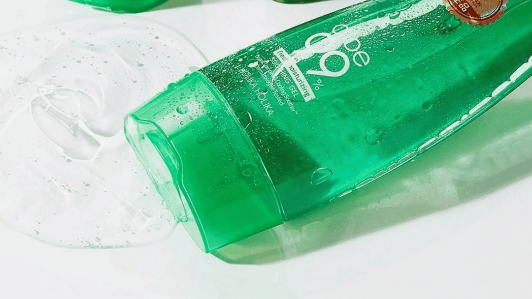 En grön tub med Holika Holika Aloe 99% Soothing Gel ligger på sidan med gelén utspridd bredvid. Tuben är delvis täckt av vatten och visar text som beskriver produktens återfuktande och lugnande egenskaper.