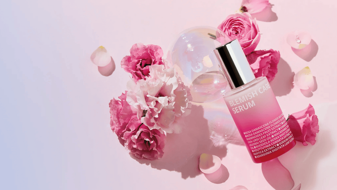 En flaska Blemish Care Serum ligger på en rosa bakgrund omgiven av rosa och vita blommor samt rosenblad. Flaskan har en silverfärgad kork och ett övertonat rosa innehåll, med produktinformation tryckt i vitt på framsidan.