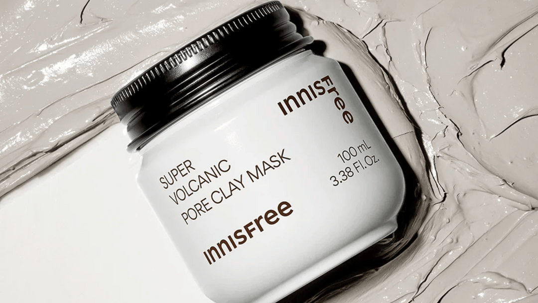 En burk av Innisfree Super Volcanic Pore Clay Mask, omgiven av den tjocka, grå lermasken som är utspridd i bakgrunden.